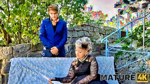 Mature silver fox cougar in black lace dress sits teasingly on outdoor bench with young suited stud behind her