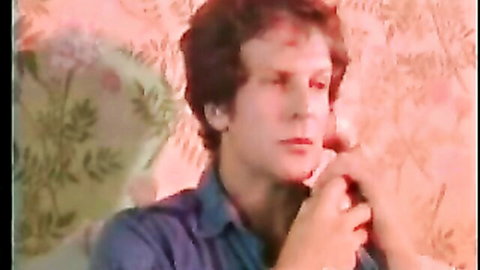 Curly-haired man in blue shirt touches lips thoughtfully against retro floral wallpaper