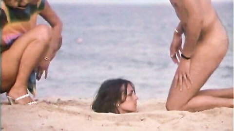 Nude man with exposed cock squats over woman's face buried in beach sand as friend watches