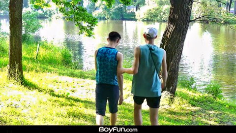 Two fit gay twinks in tight shorts hold hands walking lakeside outdoors teasing their asses