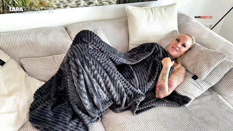 Tattooed blonde babe with big tits lounging seductively on couch wrapped in gray blanket