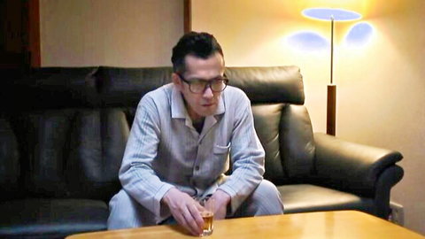 Japanese man in striped pajamas sits on leather couch holding whiskey glass pensively