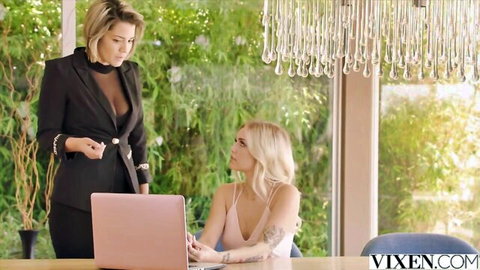 Blonde boss in sheer black top stands over sexy employee typing on pink laptop in office