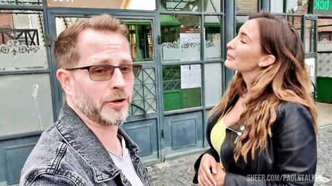Bearded man in denim jacket talks to brunette woman in yellow top and leather jacket outside graffiti wall