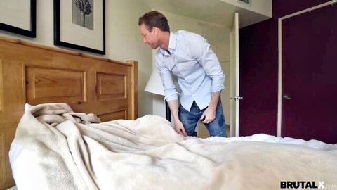 Fit man in shirt and jeans bends over bed in bedroom for Riley Star scene