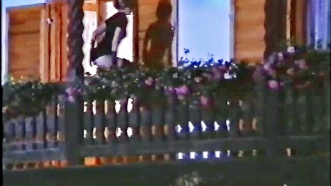 Vintage woman in black top leaning over flower-filled balcony railing teasing from behind at night