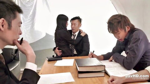 Japanese MILF in suit embraced by suited man while surrounded by businessmen at office desk in JAV threesome tease