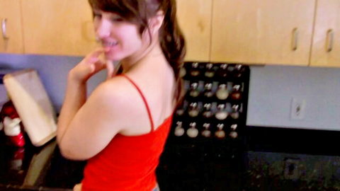 Petite brunette teen smiles seductively over shoulder in kitchen wearing tight red tank top and short skirt