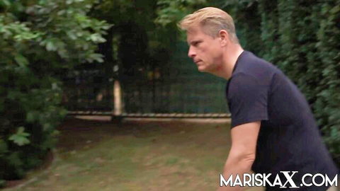 Blonde man in black t-shirt stands profile view outdoors near green fence in adult scene