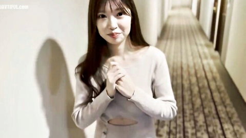 Cute Asian babe in tight gray sweater clasps hands smiling teasingly in hotel hallway