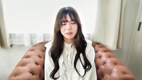 Sexy Asian woman with bangs closes eyes seductively sitting on brown leather couch in frilly white blouse