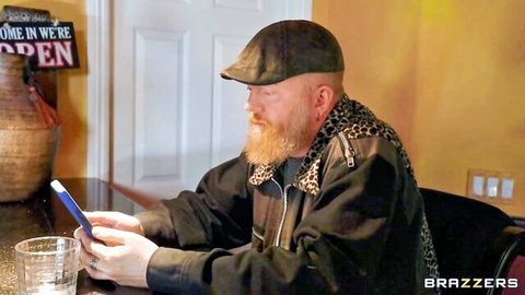Bearded bald man in flat cap and leopard scarf holds phone at kitchen table in Brazzers scene