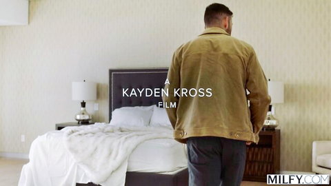 Man in beige jacket stands facing bed in Kayden Kross MILFY bedroom scene