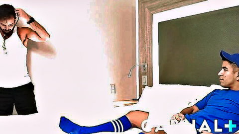 Bearded hairy hunk in tank top stands over sporty guy in blue cap and knee socks on hotel bed