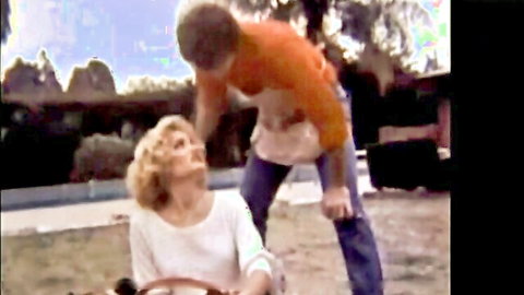 Blonde woman in white top sits outdoors as man in orange shirt bends over her intimately