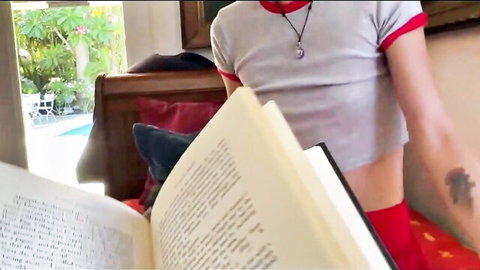 Skinny tattooed blonde in gray crop top and red shorts reading book on bed by pool