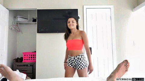 Young smiling Latina teen in neon pink tube top and checkered shorts teasing over guy on bed