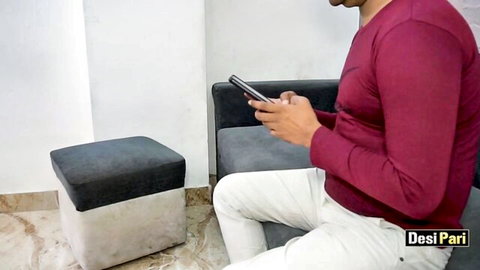 Handsome Desi Indian man in maroon shirt and white pants sits on couch holding phone teasing viewers