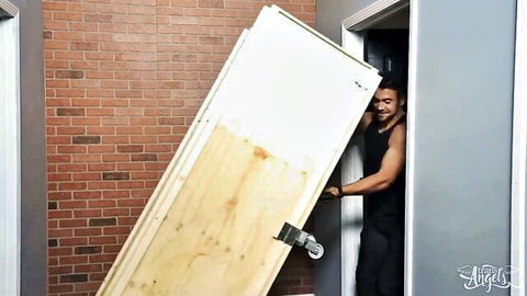 Muscular tanned man smiling while carrying large wooden door on dolly outside brick garage