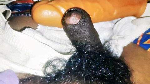 Close-up of thick erect black cock with pubes next to large orange dildo on bed sheets