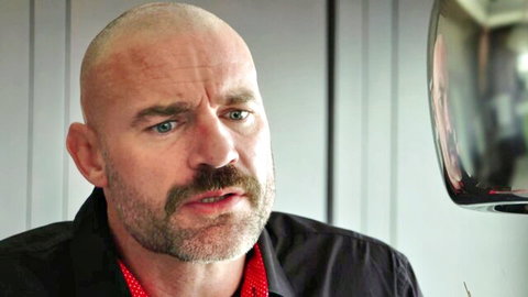 Bald man with mustache in black shirt looking sideways intensely