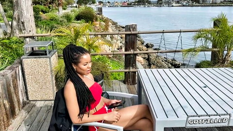 Sexy ebony babe in red bikini sitting legs spread outdoors by marina smiling