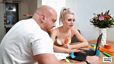 Skinny young blonde with small tits holds orange dildo as bald muscular guy watches at desk
