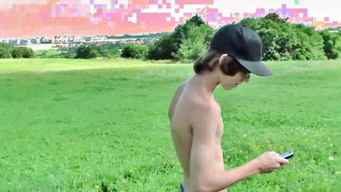 Cute shirtless college boy Adrian Leto outdoors in field, wearing cap, checking phone