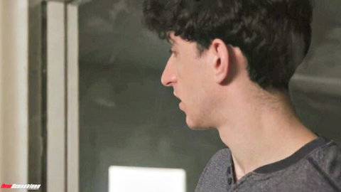 Curly-haired young man in profile view smiling slightly while wearing a gray V-neck shirt