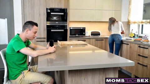 Blonde MILF stepmom in tight jeans showing round ass in kitchen while stepson sits on phone