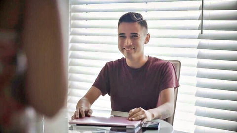 Smiling young man in maroon shirt sitting at desk with pink laptop through window blinds