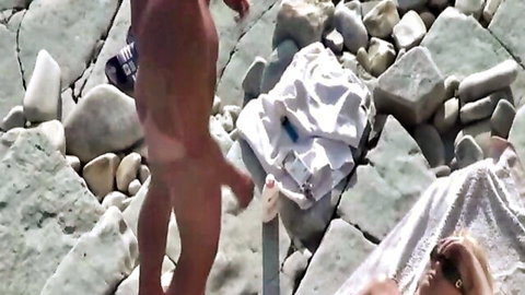 Nude blonde babe lying spread on rocky beach towel as naked guy stands with dangling cock nearby