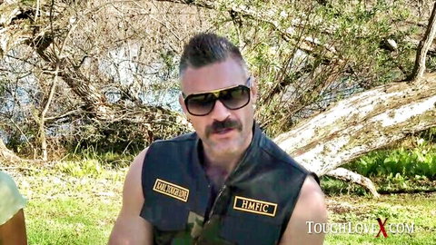 Muscular biker man with mustache and sunglasses posing outdoors in HMMC vest on ToughLove