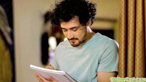 Curly haired brunette man in light blue t-shirt writing in notebook indoors