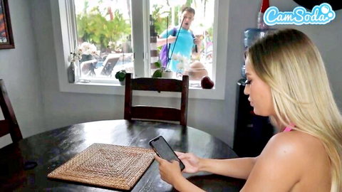 Busty blonde in pink bikini top scrolls phone at table while guy peeks from pool window