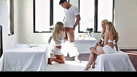 Blonde babe in lace top kneels touching standing guy's exposed hard cock while other blonde spreads legs in micro skirt threesome