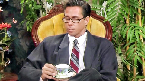 Suited man in glasses sips tea from cup in ornate yellow armchair with plants and flowers
