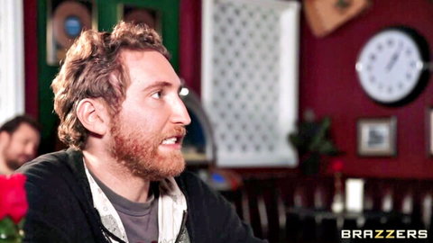 Bearded curly-haired man smiles sideways in cozy bar lounge with Brazzers logo