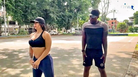 Sexy Latina with huge tits in black sports bra stands with black stud in shorts on outdoor basketball court