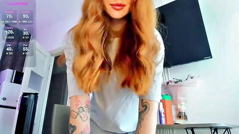 Tattooed redhead webcam girl with wavy hair smiling in loose white top teasing viewers