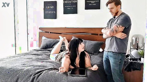 Curvy brunette lies on bed with legs up holding tablet as tattooed redhead guy watches hungrily