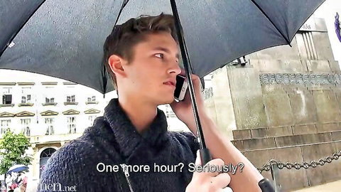 Frustrated young man in sweater talks on phone under umbrella in rain, one more hour seriously