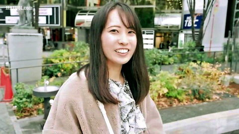 Smiling Asian woman in beige cardigan and blouse standing outdoors in urban park