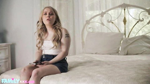 Petite blonde teen in tight tank top and shorts sits on bed holding phone with shocked face