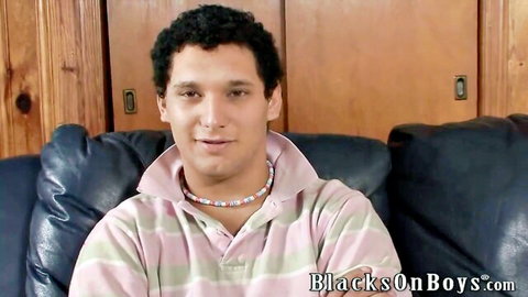 Handsome curly-haired guy in pink polo shirt and beaded necklace sits smiling on couch for gay interracial fun