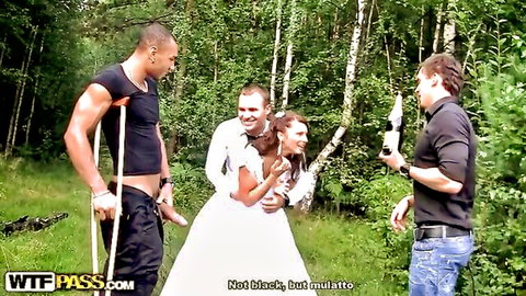 Bride in wedding dress embraced by groom in woods with two men watching