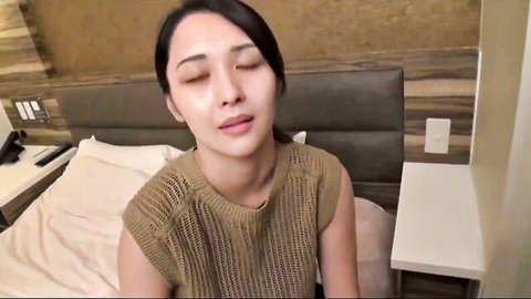 Sexy Asian woman in tight beige sleeveless top smiling seductively on hotel bed POV