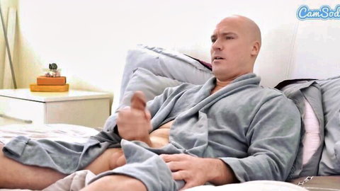 Bald muscular man in open gray robe exposes his thick cock and balls while gesturing on bed