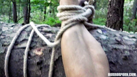 Fair-skinned wrist tightly bound with thick rope to mossy tree log in dark forest BDSM outdoor