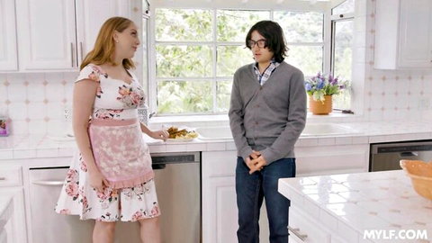 Sexy redhead MILF in floral dress and apron serves pancakes to shy young guy in kitchen